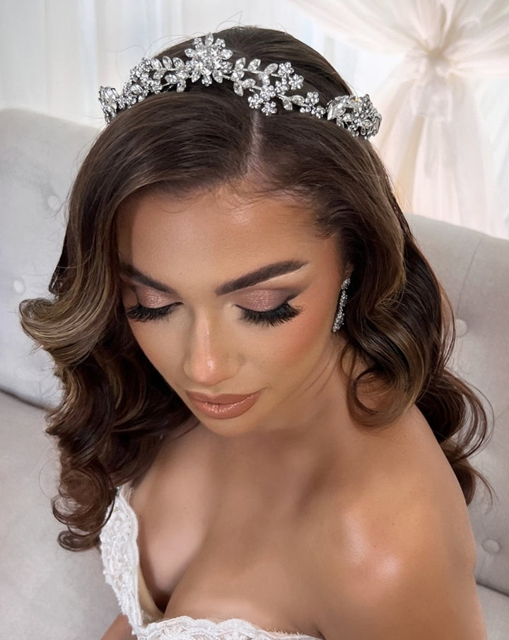 A woman with long, wavy dark hair wears a Delicate Floral Tiara style no. 80008 and elegant earrings. Accented by a Swarovski tiara, she has soft makeup with long eyelashes and glowing skin. Dressed in a strapless white gown, shes seated on a light-colored, tufted chair.