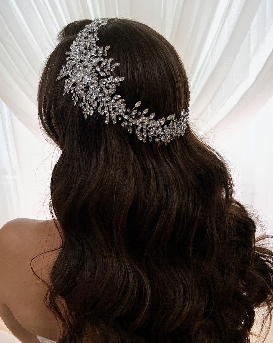 A woman with long, wavy brown hair is seen from behind, adorned with the Dramatic Crystal Bridal Halo style no. 242518. Shes wearing an off-shoulder dress, and a sheer white curtain is in the background.
