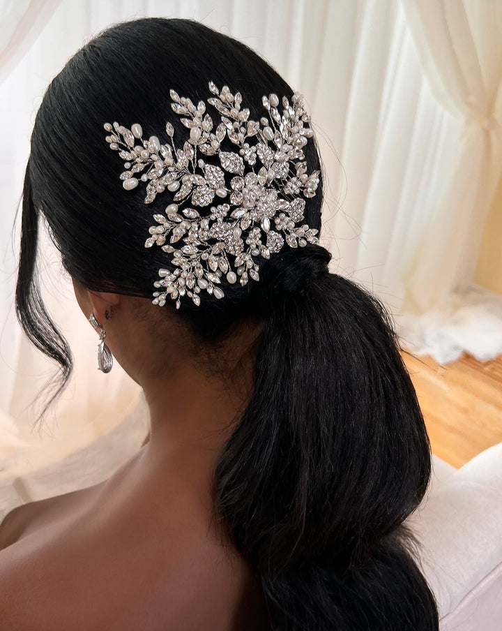 A woman with long dark hair styled in a low ponytail displays a Floral Crystal and Pearl Hair Comb (style no. 242716) positioned above the ponytail. She is adorned with drop CZ bridal earrings, sitting against a background of white draped fabric.