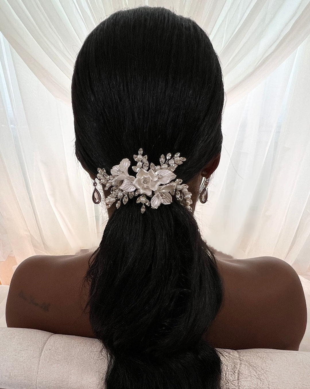 A person with long black hair is seen from behind, styled in a low ponytail, adorned with the Delicate Blossoming Swarovski Crystal Bridal Comb (style no. 242733), featuring pearls and crystals. Sheer white curtains are softly draped in the background.