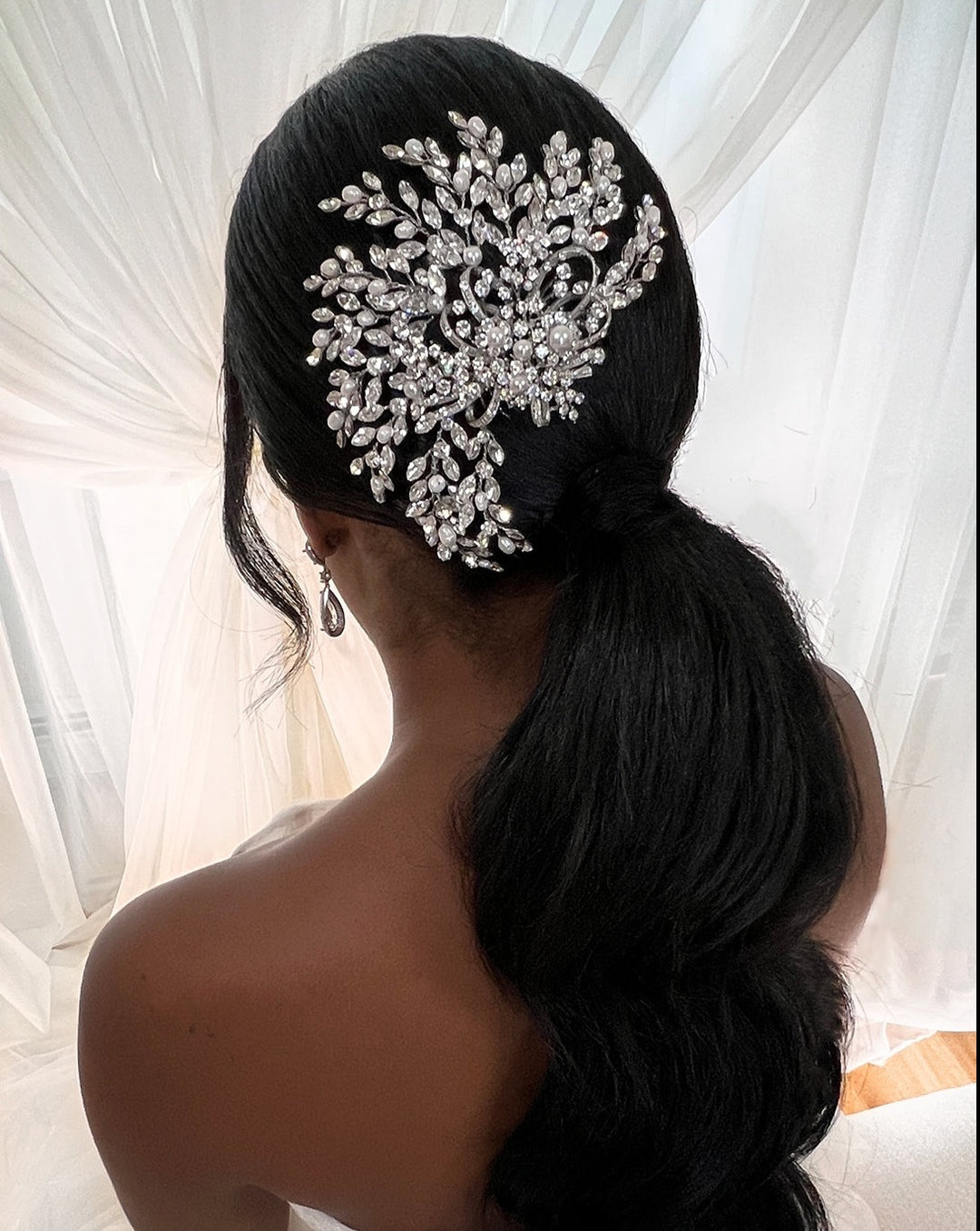 A person with long, dark hair styled in a low ponytail adorned with the Swirling Pearl and Crystal Comb (style no. 242580) featuring floral motifs. A soft, light background shows sheer curtains partially visible.