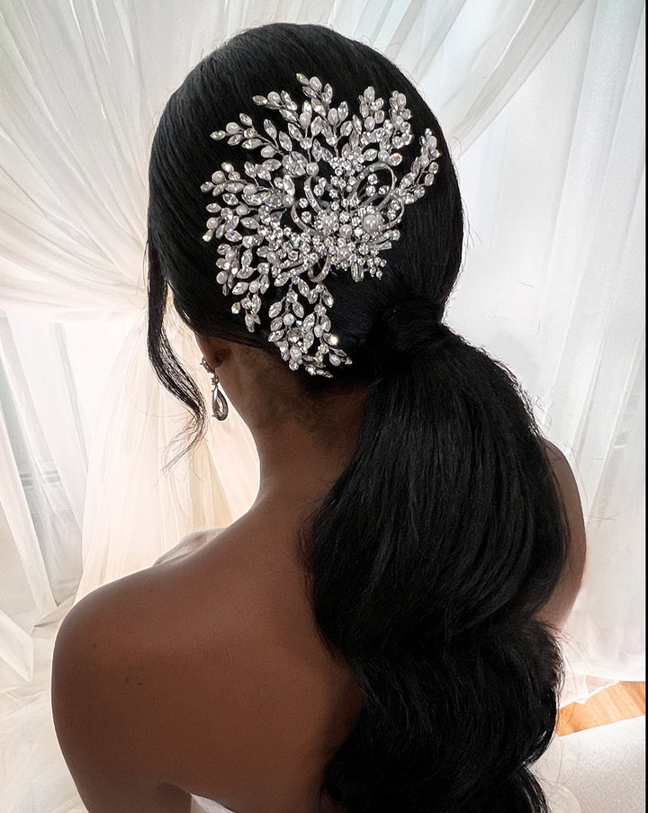 A person with long, dark hair styled in a low ponytail adorned with the Swirling Pearl and Crystal Comb (style no. 242580) featuring floral motifs. A soft, light background shows sheer curtains partially visible.