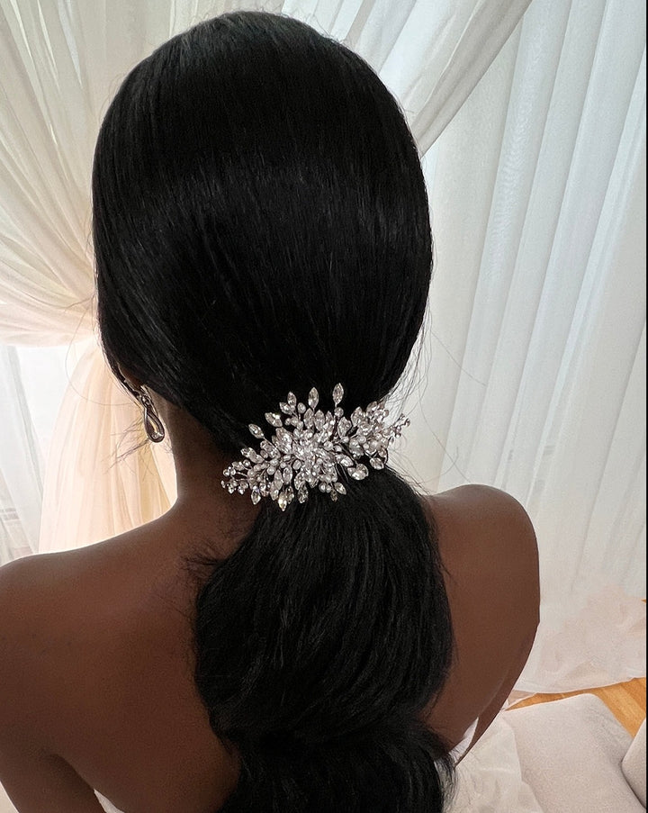 A person with long dark hair styled in a low ponytail is adorned with the Dainty Crystal Comb (style no. 242731) featuring sparkling Swarovski crystals. The setting is softly lit, with sheer white curtains in the background.