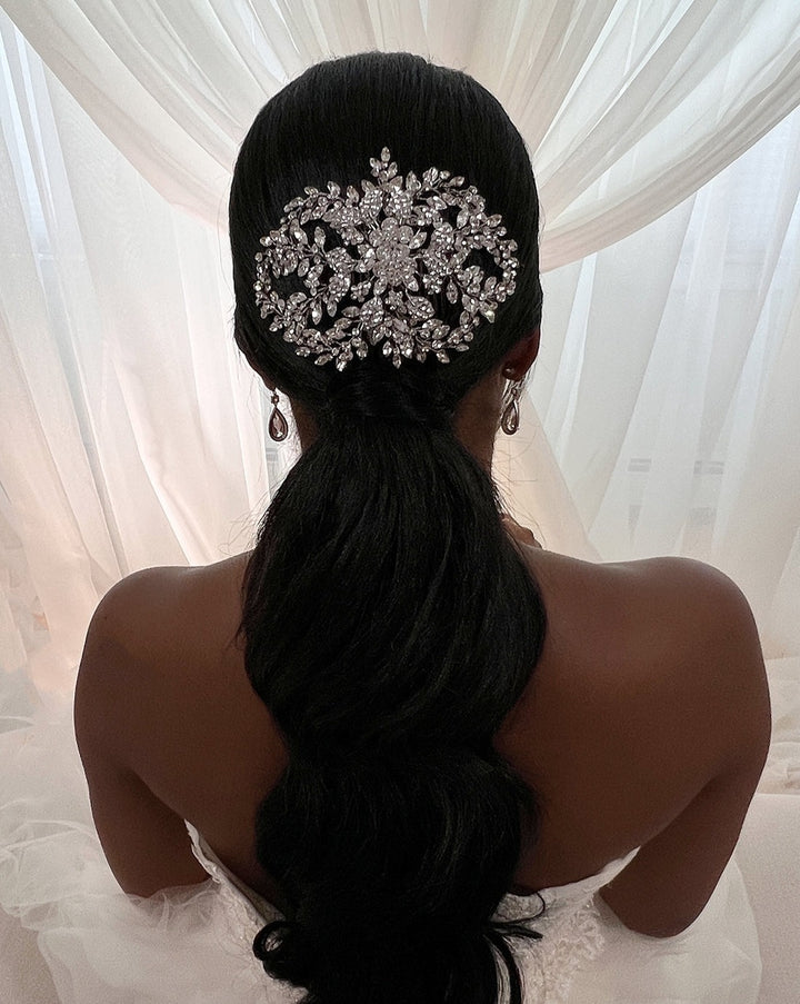 A woman with dark hair styled in a low ponytail sits gracefully in front of sheer white curtains, her look elevated by the Crystal Bloom Swarovski Crystal Hair Comb style no. 242717. She wears a white outfit with thin straps, perfectly complementing the intricate sparkle of her bridal headpiece.