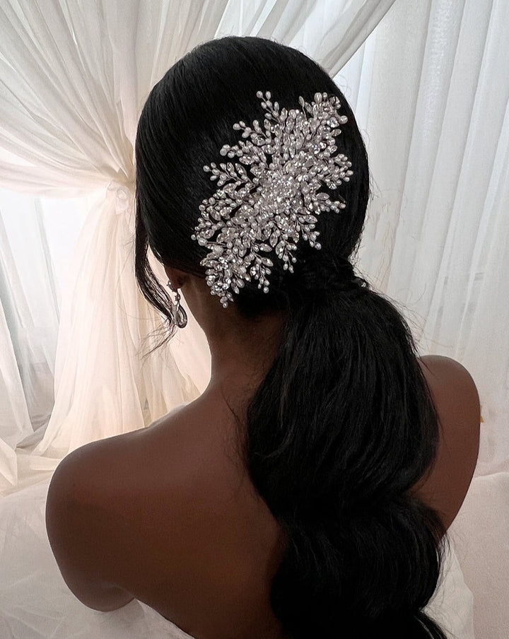 Rear view of a person with long, dark hair in a low ponytail adorned with the Classic Swarovski Crystal Bridal Comb (style no. 242646) resembling a snowflake. They are wearing CZ wedding earrings and sitting in front of flowing white drapery.