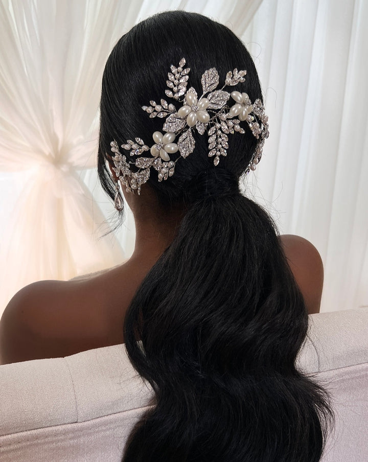 A person with long, wavy black hair styled in a low ponytail, adorned with the intricate Branching Pearl and Crystal Bridal Hair Comb (style no. 80027) featuring leaf and floral designs, is seated on a light-colored sofa with sheer white curtains in the background.