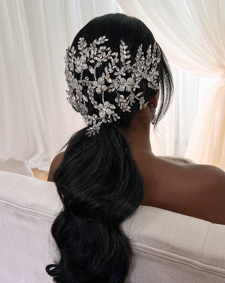 A person with long, wavy black hair sits facing away, their low ponytail adorned with an ornate, silver floral hairpiece reminiscent of the sparkling Dramatic Crystal Headband (style no. 242695). Soft white curtains illuminate the background.
