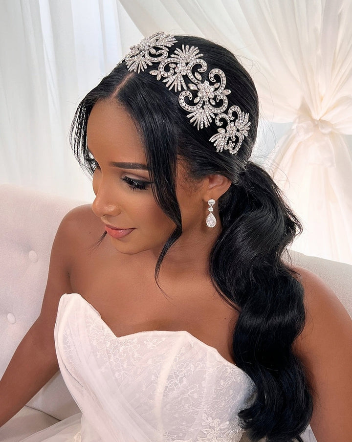 A bride wearing a white strapless wedding dress and the Showstopper Crystal Headband (Style no. 3913542) sits on a white cushioned seat. Her long, dark hair cascades over her shoulder. Teardrop earrings enhance her look, with a softly draped background completing the scene.