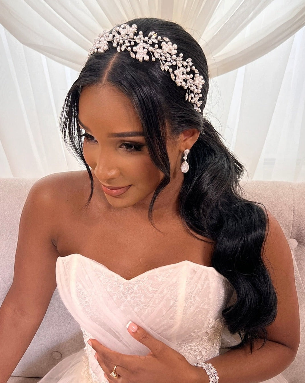 The bride, in a strapless white lace gown, sits against a soft curtain backdrop. Her look is elevated by a floral Swarovski crystal headband and perfectly manicured nails.