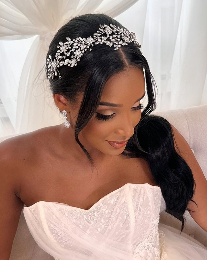 A bride with long black hair and elegant makeup sits on a couch in a white, strapless lace wedding gown. She wears the  Delicate Weaving Swarovski Crystal Vine tiara (style no. 242465). Her hair is styled in a low ponytail, complemented by drop CZ wedding earrings.