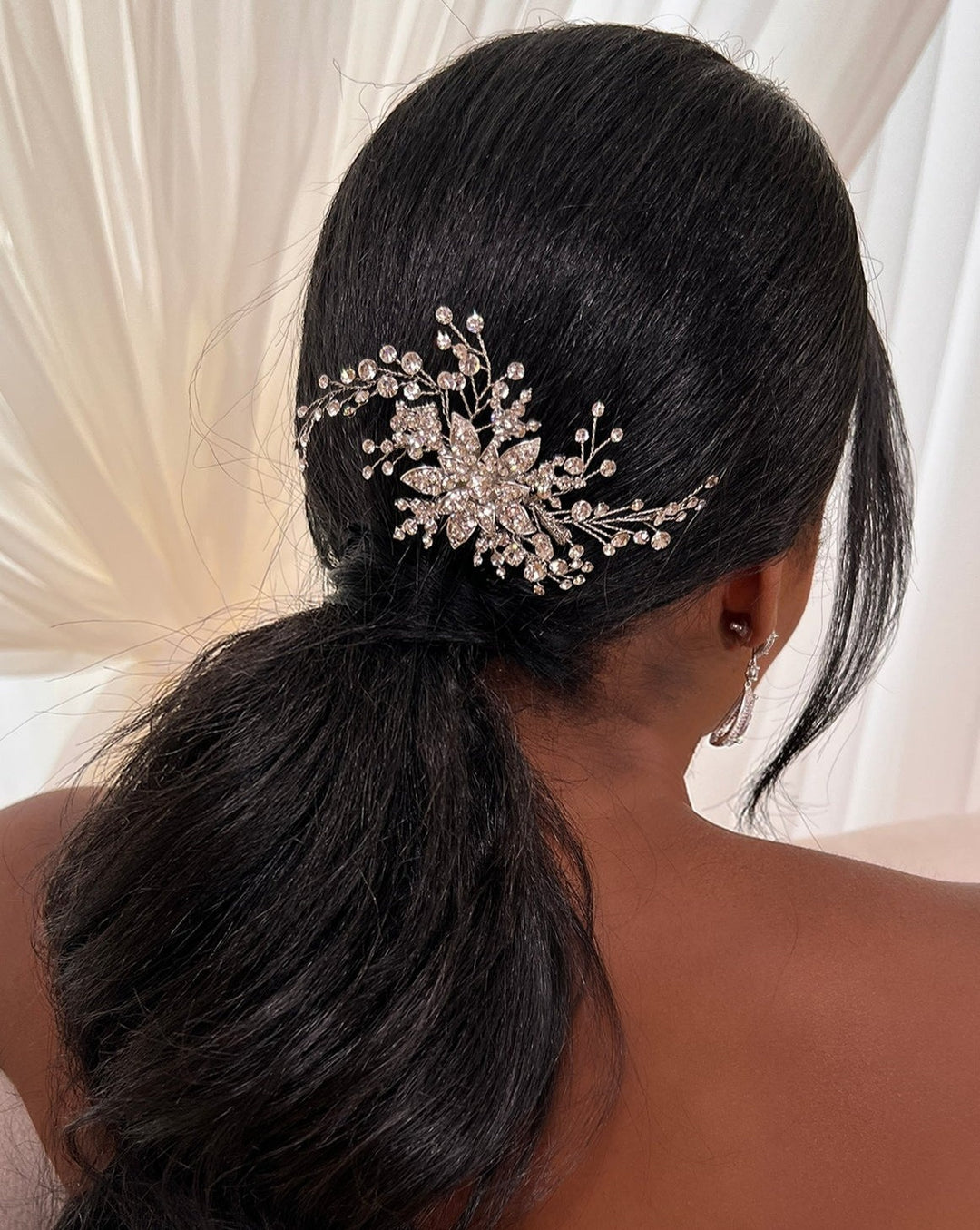 A woman with long black hair in a low ponytail wears a Delicate Flower Swarovski Crystal Hair Comb, style no. 242697, adorned with floral and branch designs. Her look is completed by elegant CZ wedding earrings and soft draped fabric in the background.