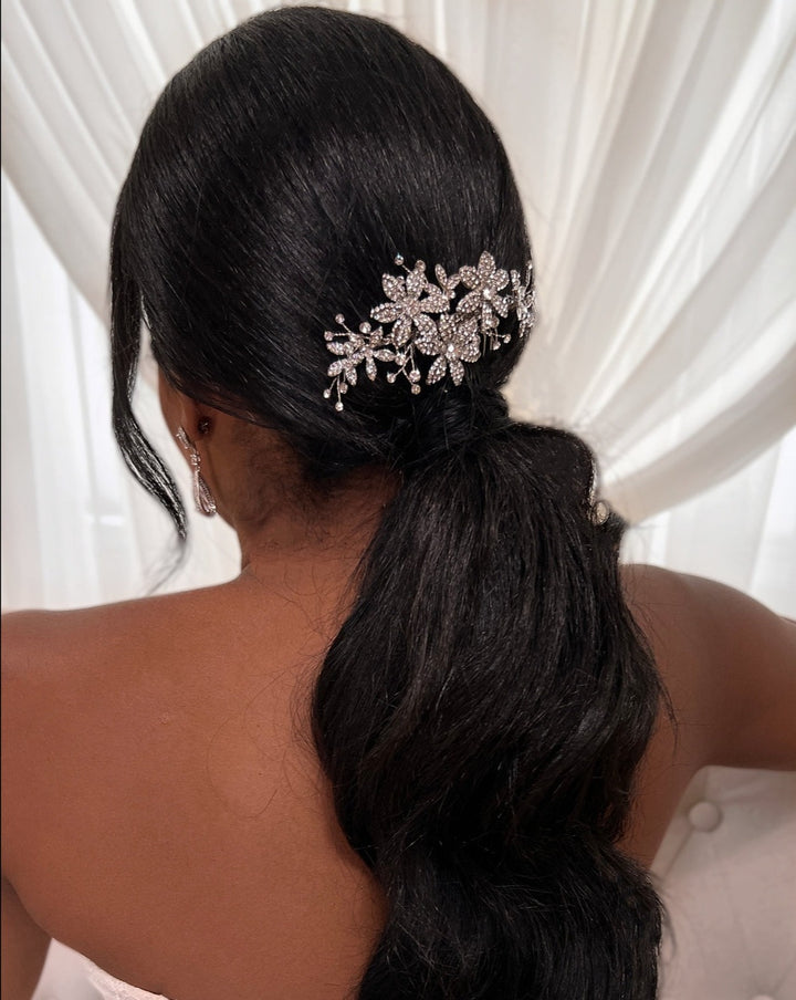 A woman with long black hair styled in a low ponytail showcases the Delicate Floral Comb style no. 242692, against a white curtain backdrop.