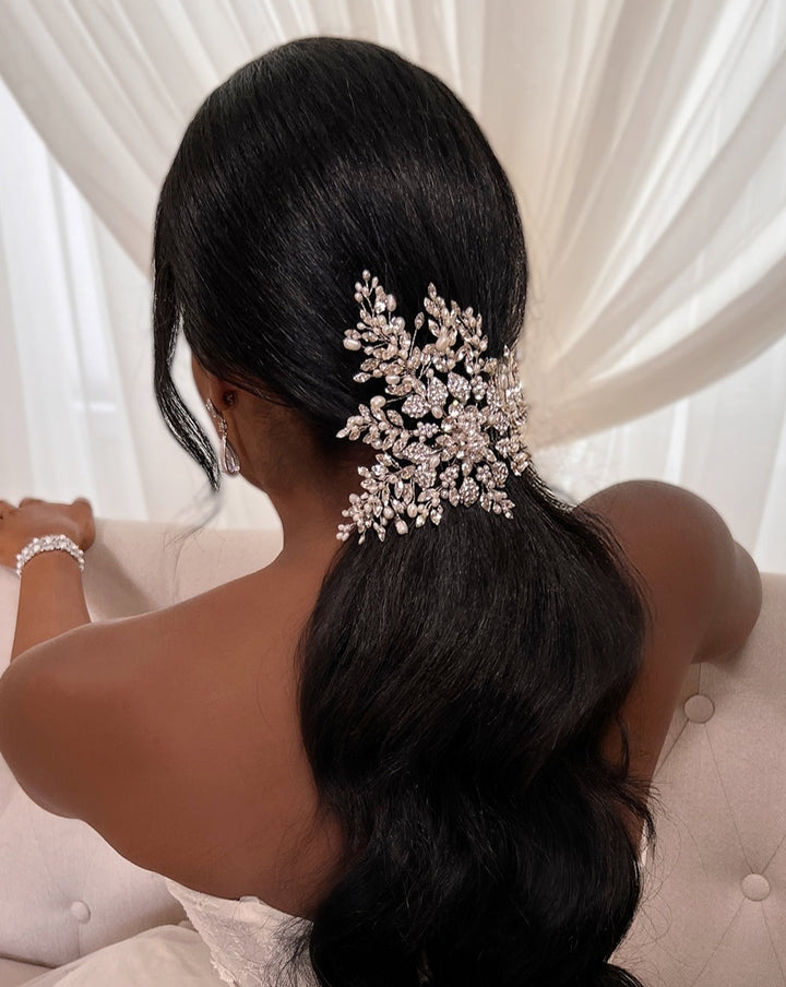 A woman with long, dark hair styled in a loose ponytail, adorned with the Floral Crystal and Pearl Hair Comb (style no. 242716), sits on a light-colored couch. She wears a strapless white dress and earrings, with sheer curtains in the background.