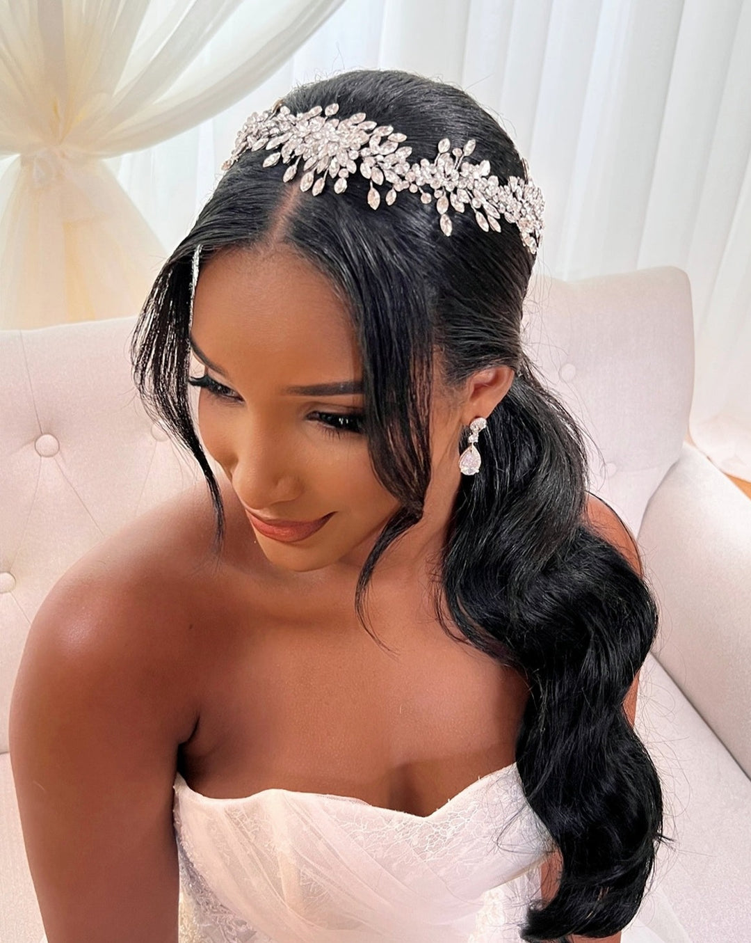 The bride with long, wavy hair sits on a white sofa, adorned with the Shimmering Swarovski Crystal Bridal Halo style no. 242542 that glimmers in the light. Her crystal  headpiece and earrings frame her serene expression, while her strapless white dress complements the softly lit backdrop.