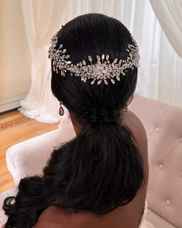 A woman with long dark hair styled in a low ponytail adorned with the Shimmering Crystal Halo (style no. 242542), sits on a light pink chair, sheer white curtains softly framing the backdrop.