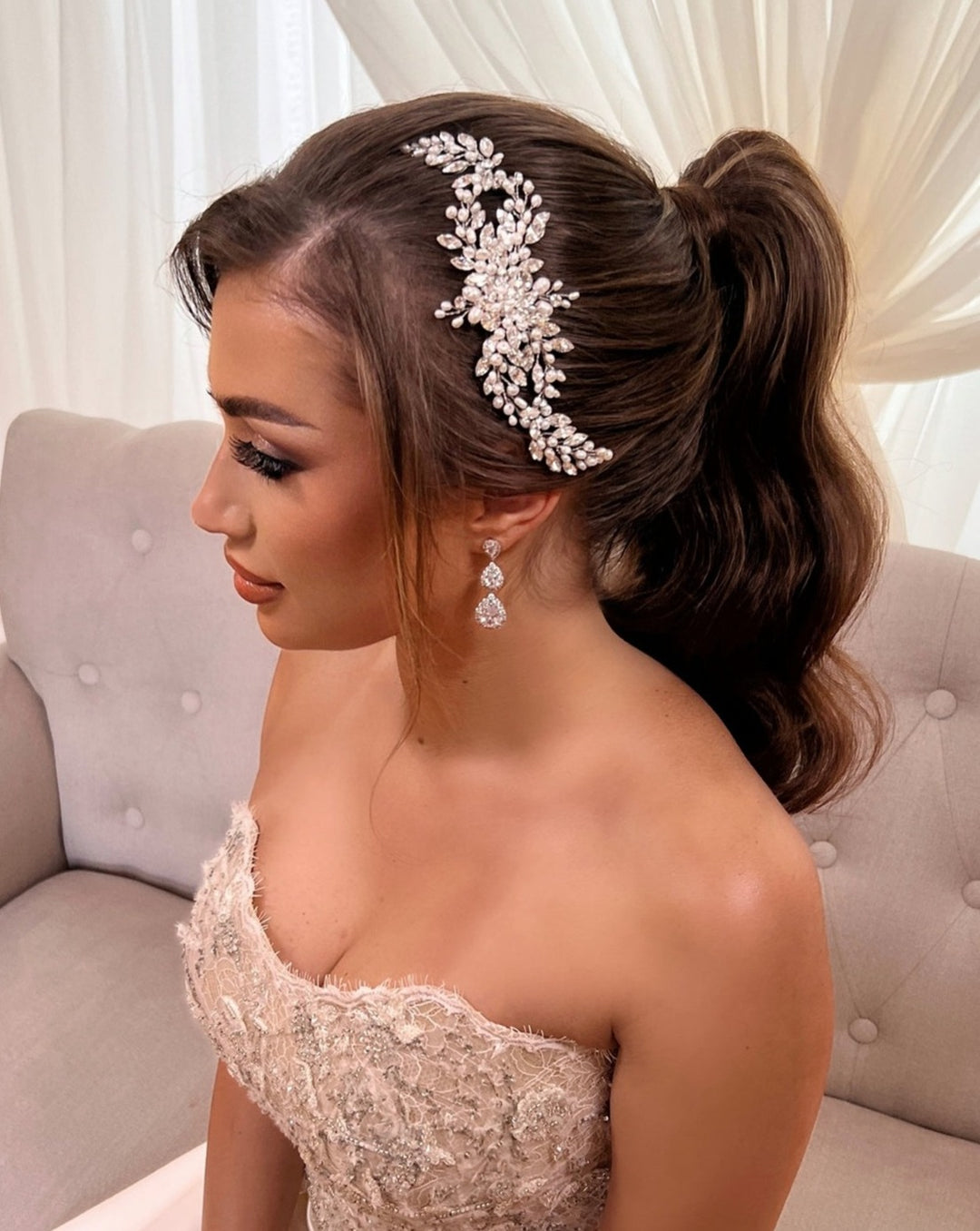 A woman in a strapless lace dress sits on a sofa, showcasing her side profile. Her hair is styled in a high ponytail adorned with the Flowing Cluster Hair Comb style no. 242514, featuring intricate silver and pearl details, and she wears elegant drop earrings completing her refined look.