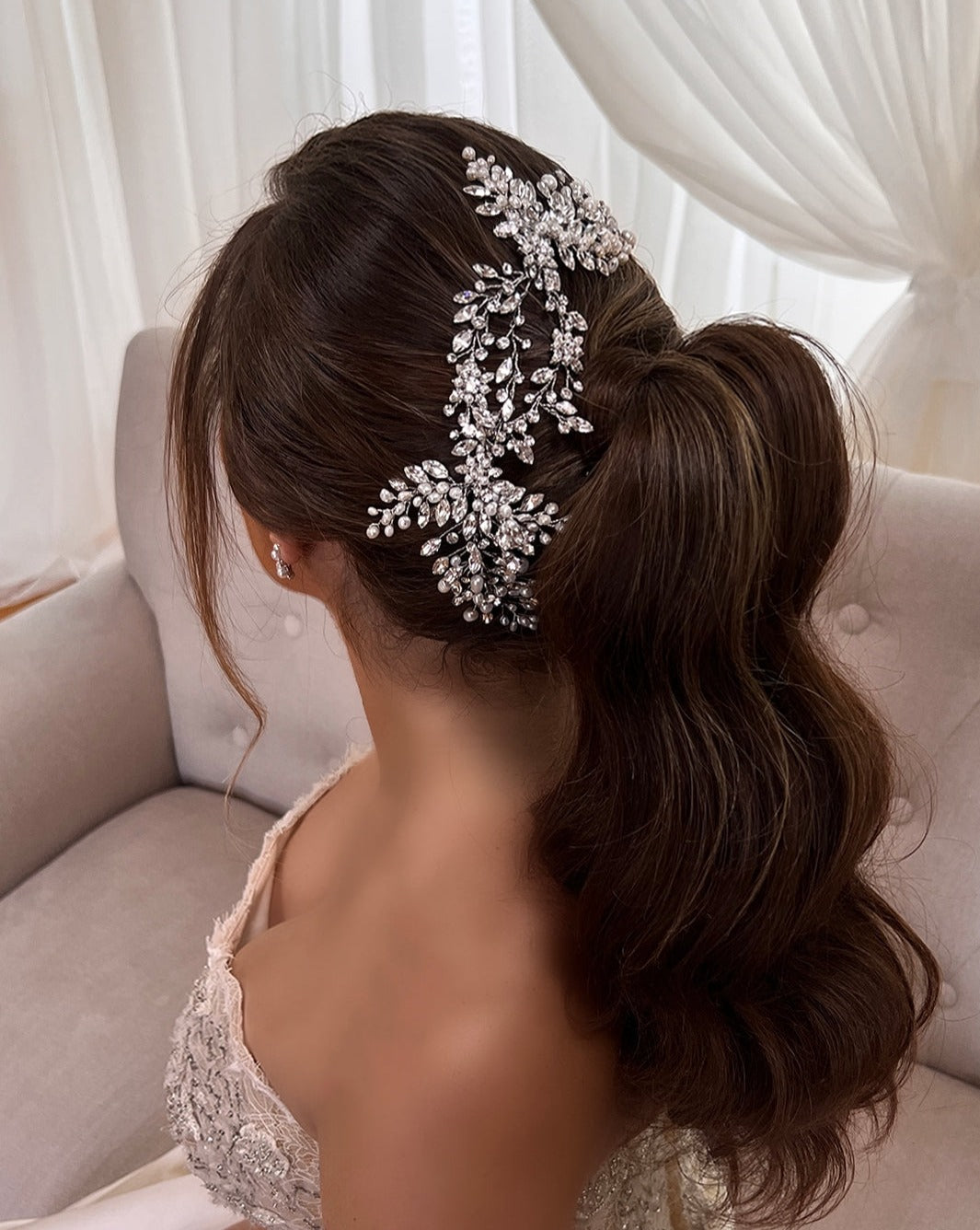 A woman with wavy brown hair in a low ponytail, adorned with an intricate Swarovski crystal floral bridal headpiece sits on a beige couch with sheer curtains. She is wearing a cream lace dress.