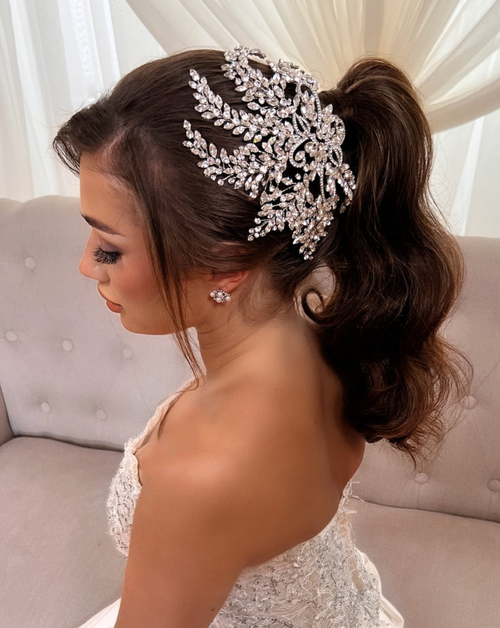 A woman in a strapless lace dress with wavy hair in a ponytail, adorned with the Glamorous Crystal Bridal Comb style no. 242535 and matching CZ wedding earrings, sits on a beige sofa with a white curtain backdrop.