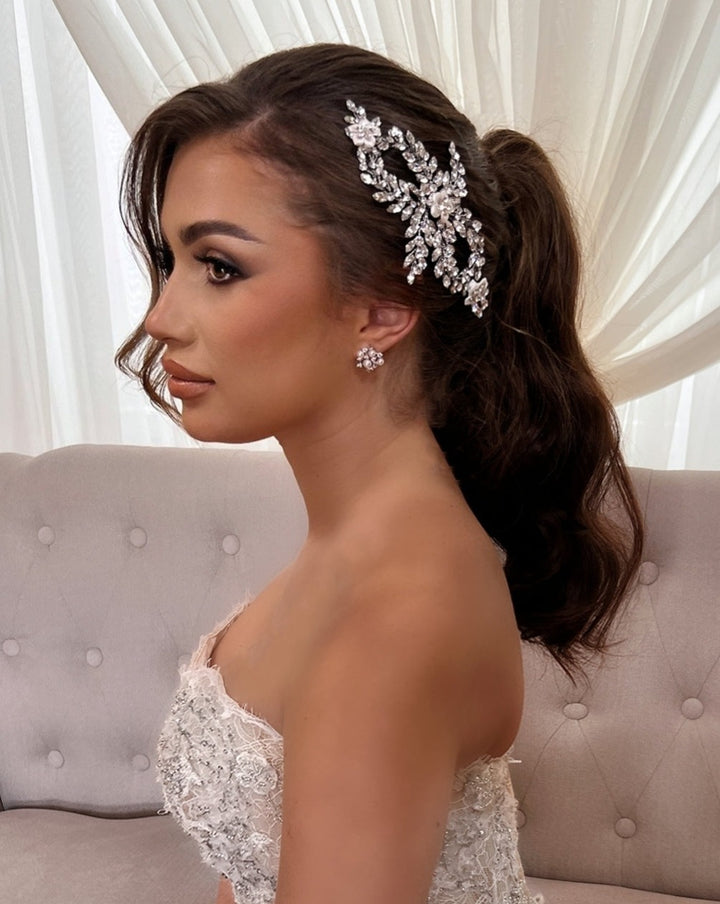 A woman in a strapless lace dress sits on a tufted sofa, her long hair styled in a low ponytail adorned with the Swarovski Crystal and Porcelain Flower Bridal Hair Comb (style no. 242726) that sparkles. She wears matching floral earrings, elegant makeup, with soft lighting and sheer curtains enhancing the ambiance.