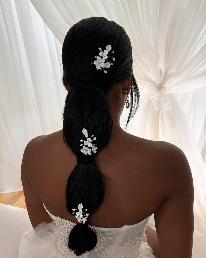 A person with long, dark hair is styled in a low segmented ponytail, adorned with elegant Porcelain Flower Pin style no. 242723 and Swarovski hair combs. Theyre wearing a white strapless dress, standing gracefully before sheer white curtains.