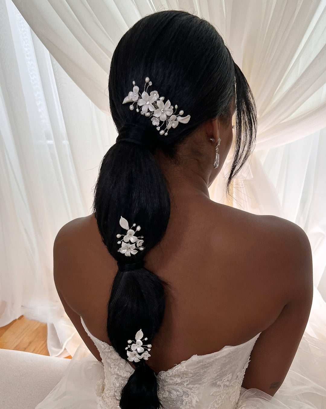 A woman with long dark hair styled in a low ponytail adorned with Romantic Porcelain Flower Hair Comb style no. 242724. She wears a strapless white lace dress, sitting gracefully before sheer white curtains, her back facing the camera.