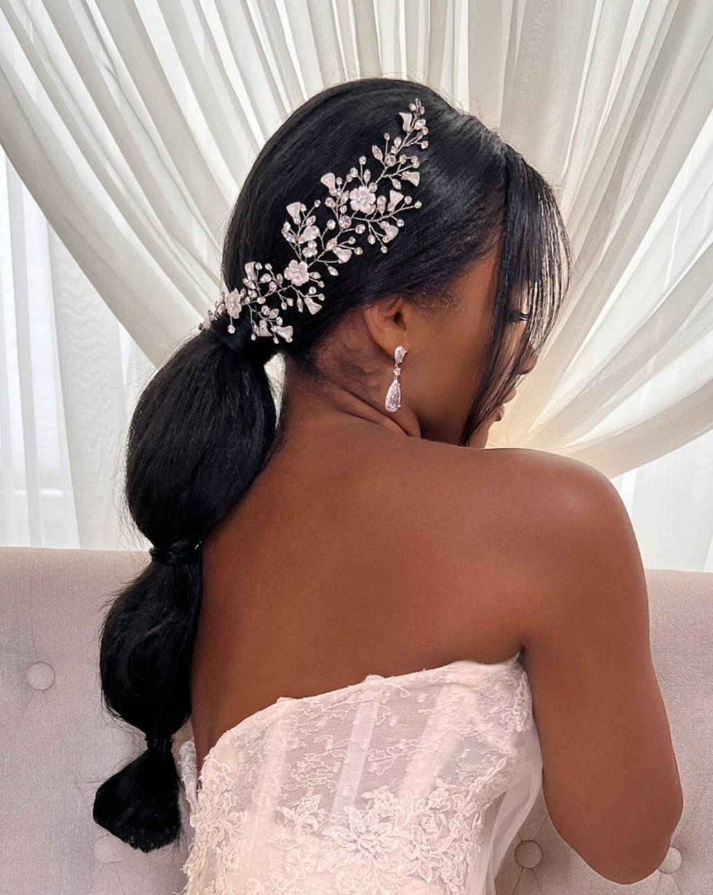 A woman in a strapless white lace dress faces away from the camera, her long black hair styled in a low bubble ponytail adorned with the Asymmetrical Blossom Swarovski Crystal Hair Vine (style no. 242735) complemented by dangling earrings against sheer curtains.