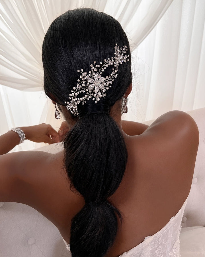 A woman with long black hair styled in a low ponytail adorned with the Swarovski Crystal Flower Vine (style no. 242707), wearing pearl bridal earrings and a white lace strapless dress, sits on a tufted chair against sheer white drapes.