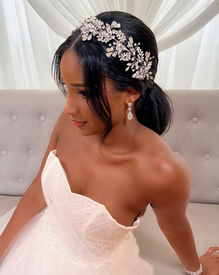 A bride in a strapless white gown sits on a light gray sofa, her hair styled in a low ponytail featuring the Teardrop Swarovski Crystal Hair Vine, style no. 242696. She wears drop earrings and gazes downward, with white drapery visible in the background.
