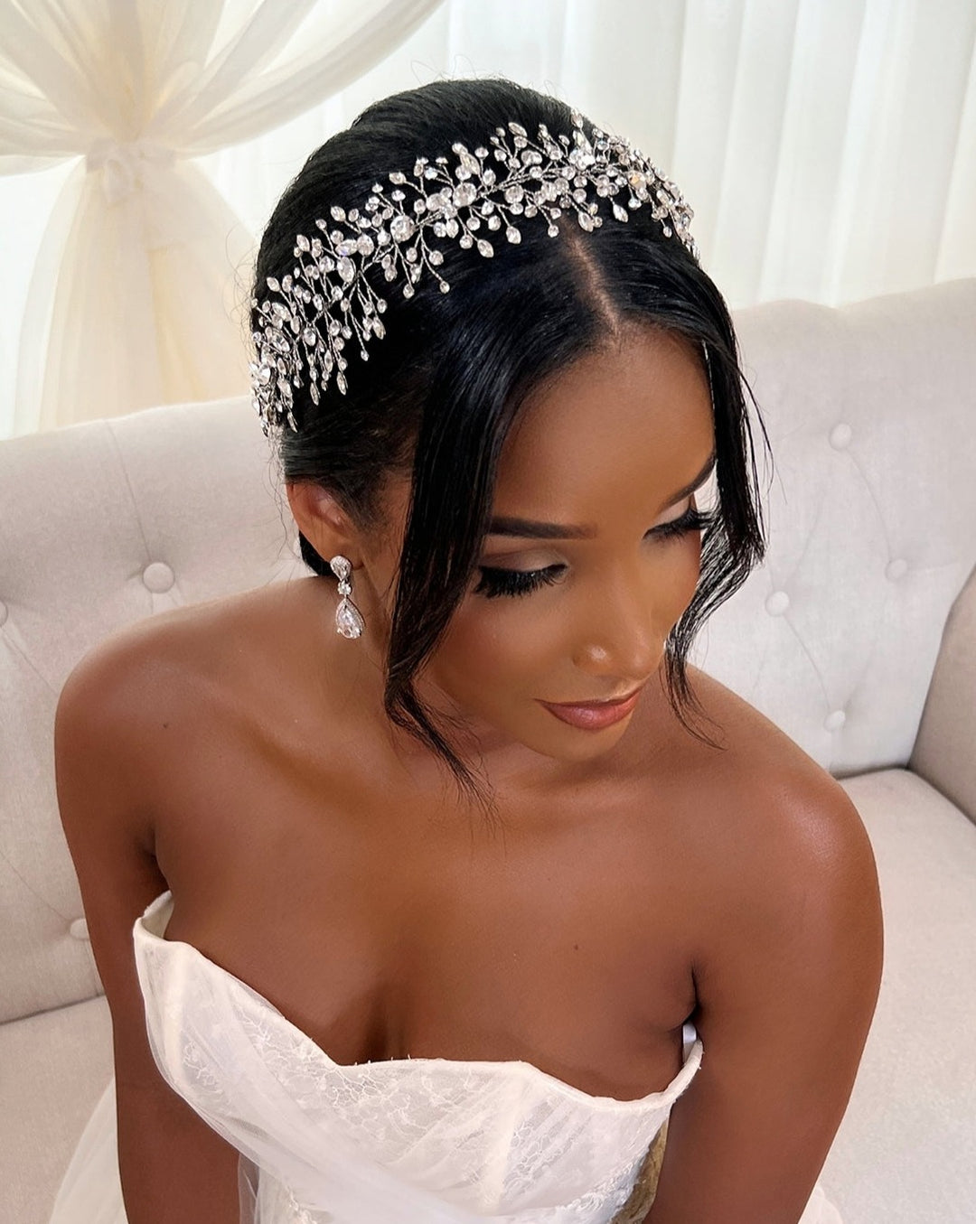 A bride sits on a light sofa in a strapless white wedding dress, her hair adorned with the Elegant Branching Crystal Vine, style no. 242699. This ornate Swarovski hair vine adds sparkle to her look. She wears drop earrings and elegant makeup, all set against sheer white curtains.