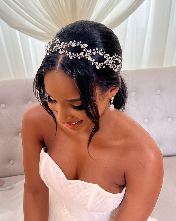 A bride sits on a light sofa, smiling while gazing down. She wears a white strapless wedding dress, Elegant Looping Bridal Vine style no. 39228878, and drop CZ wedding earrings. Her hair is in a sleek updo as soft light filters through sheer curtains behind her.