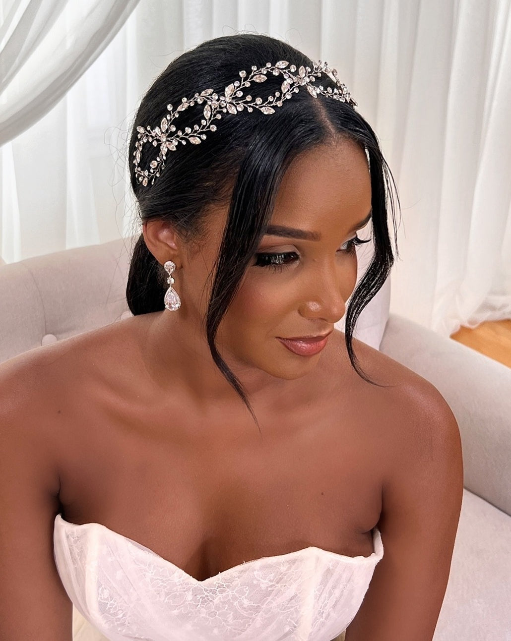 A woman in a strapless white dress sits on a light sofa, her long dark hair styled with the Elegant Looping Bridal Vine (style no. 39228878) and elegant CZ wedding earrings. In the softly lit room, she gazes sideways exuding timeless elegance.