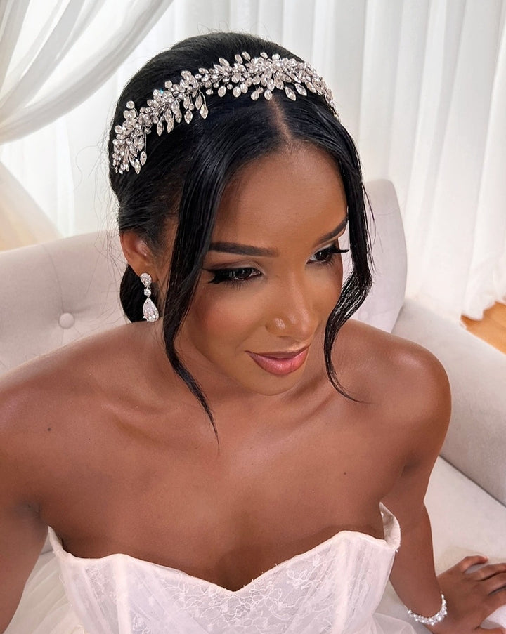 A bride sits on a light-colored sofa in a strapless white gown, her hair elegantly styled with the dazzling Elegant Swarovski Crystal Cluster Hair Vine, style no. 242517. She wears matching earrings and bracelet, with soft makeup accentuating her features against a softly lit backdrop of white curtains.