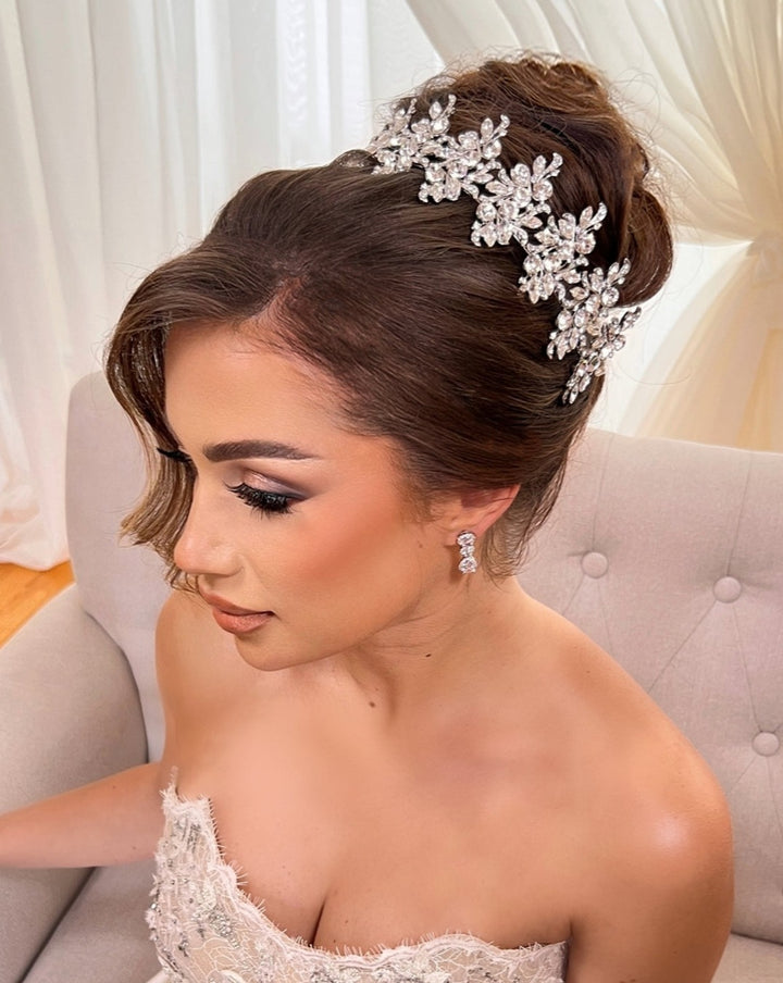 A woman in a strapless dress sits on a light gray chair, her elegant updo adorned with the Luxurious Pearl and Crystal Headband Style no.80066, complementing her subtle makeup and sparkling earrings. Light-colored curtains form the background.