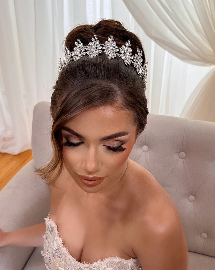 A woman with styled hair adorned with a Luxurious Pearl and Crystal Headband Style no. 80066 sits on a light chair. She wears a strapless white lace dress, her eyes closed and makeup elegant, in a softly lit room with white curtains in the background.