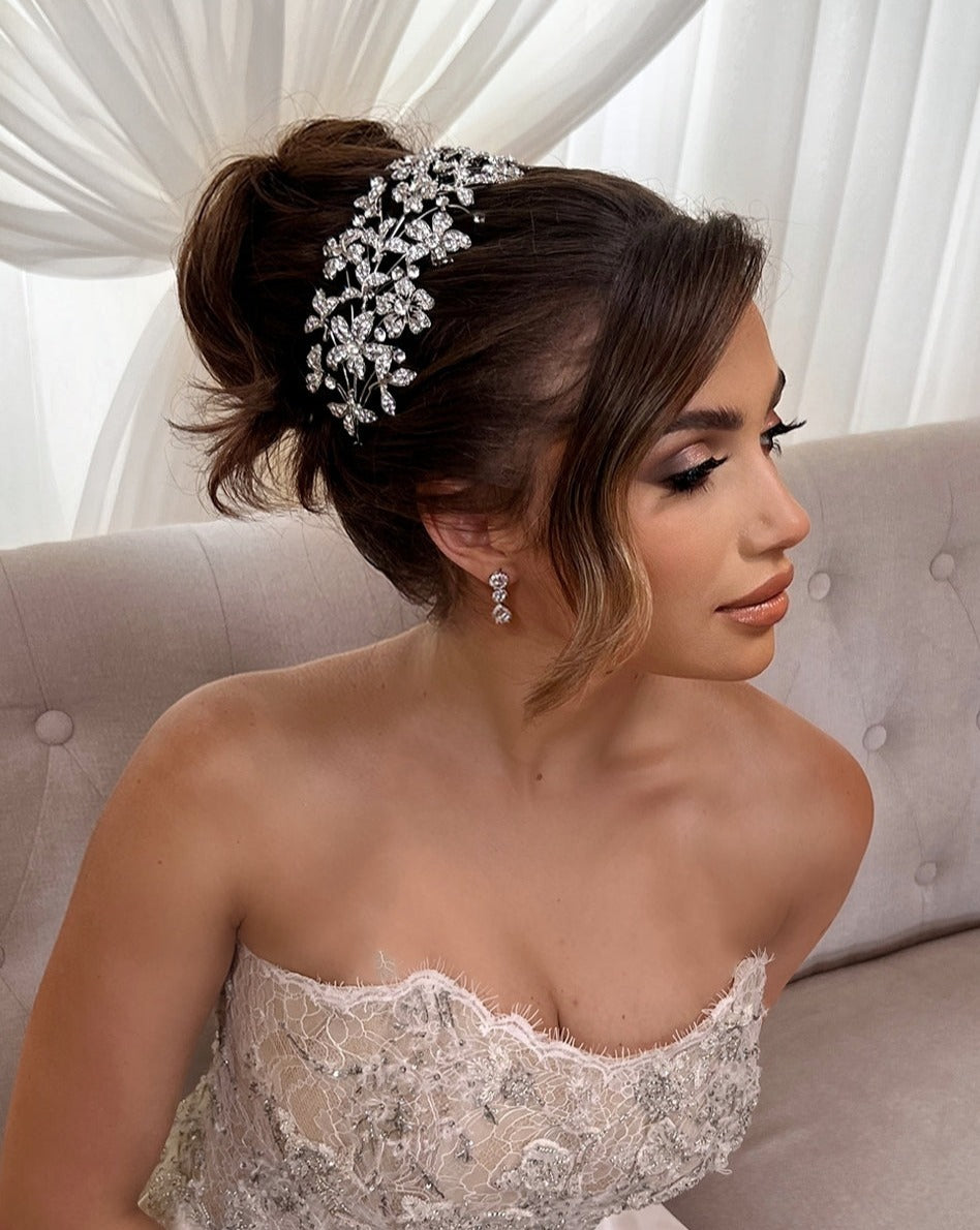 A woman in a strapless white lace gown sits on a tufted sofa. Her hair, styled in an elegant updo, is adorned with the Dazzling Crystalized Flower Headband (style no. 242678). She wears drop bridal earrings and soft, natural makeup, gazing thoughtfully to the side.