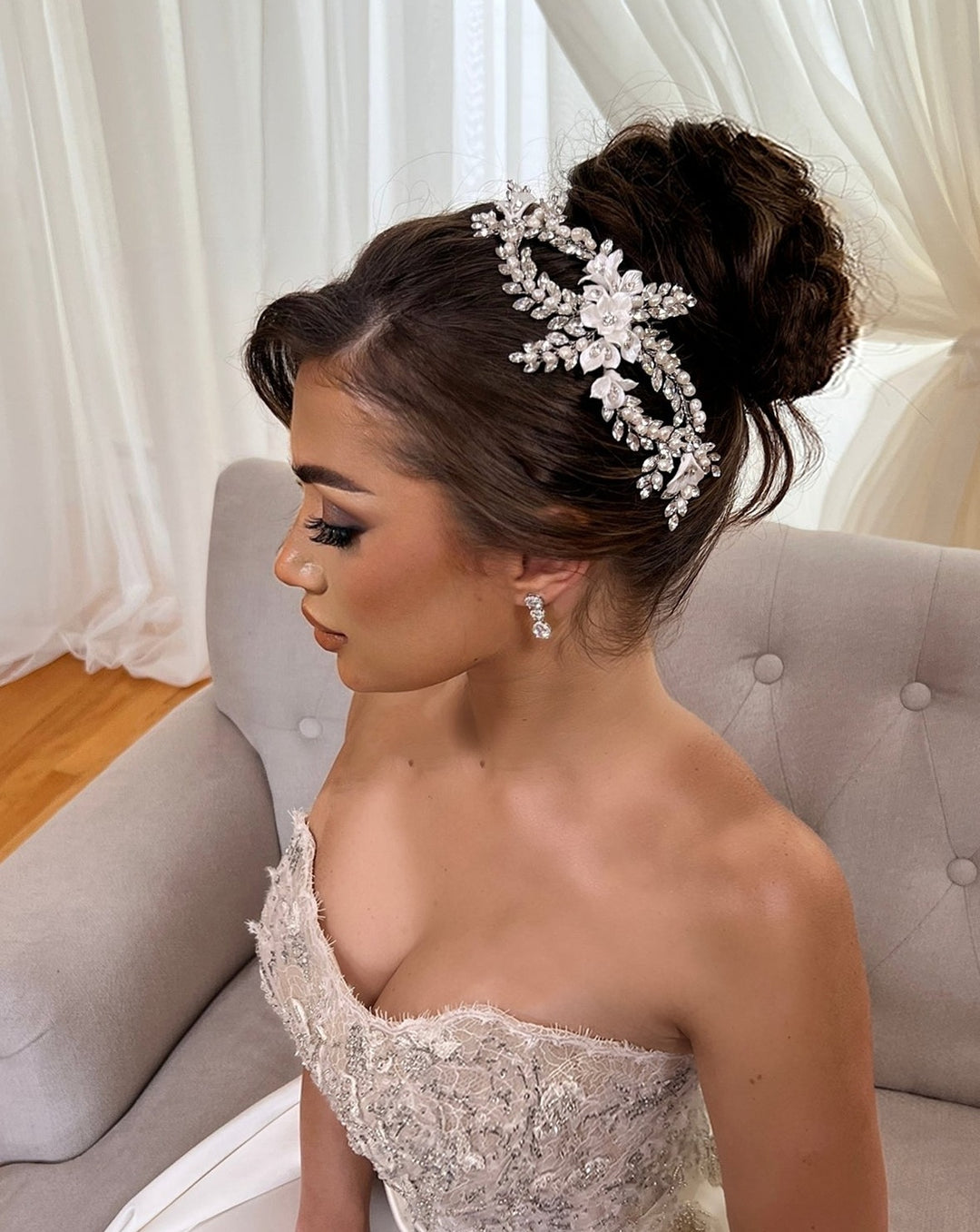 A bride sits gracefully on a couch against a light curtain. Her hair is styled in a bun, adorned with the Dazzling Pearl and Flower Bridal Hair Comb style no. 242734. Her strapless lace gown showcases intricate detailing, and her makeup is elegantly done.