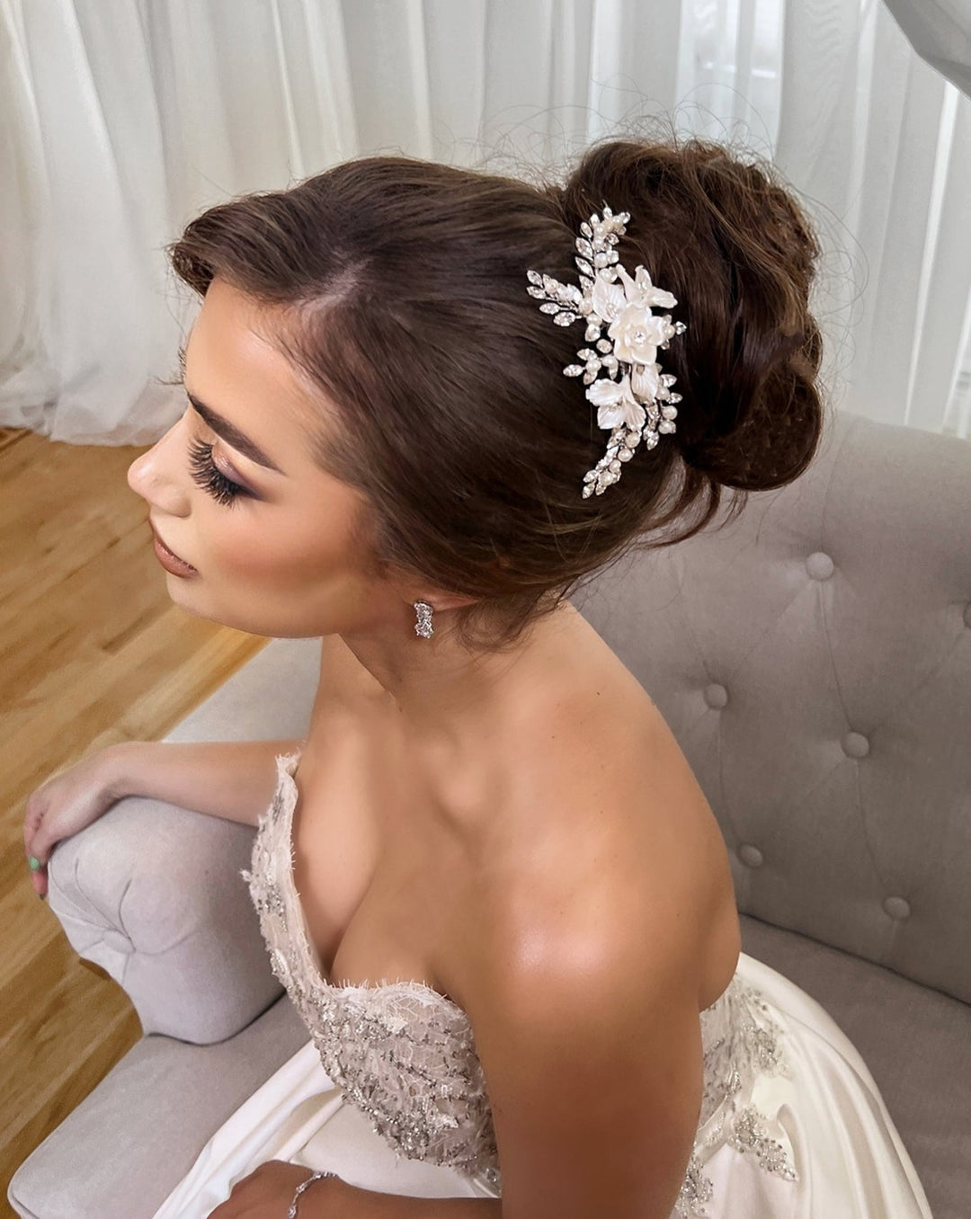 The bride, with an elegant updo adorned by a Delicate Blossoming Swarovski Crystal Bridal Hair Comb (style no. 242733), sits gracefully on a light chair. She wears a strapless lace gown and stud earrings, surrounded by softly lit interiors with sheer curtains, her eyes closed to showcase her natural makeup.