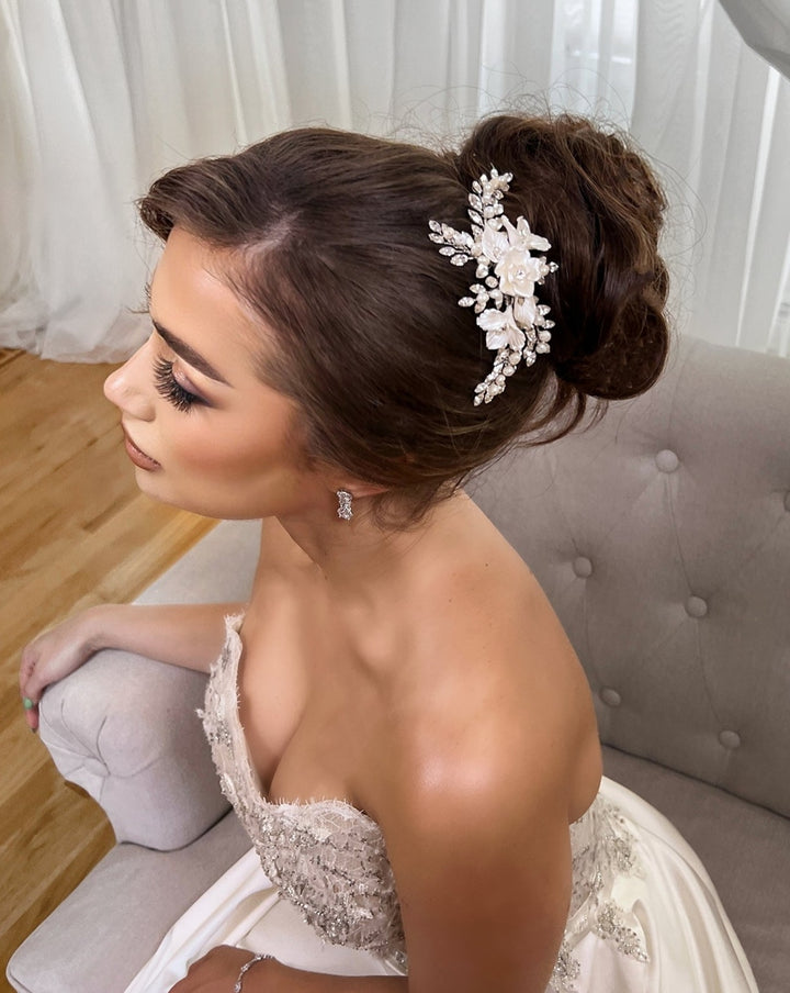 The bride, with an elegant updo adorned by a Delicate Blossoming Swarovski Crystal Bridal Hair Comb (style no. 242733), sits gracefully on a light chair. She wears a strapless lace gown and stud earrings, surrounded by softly lit interiors with sheer curtains, her eyes closed to showcase her natural makeup.