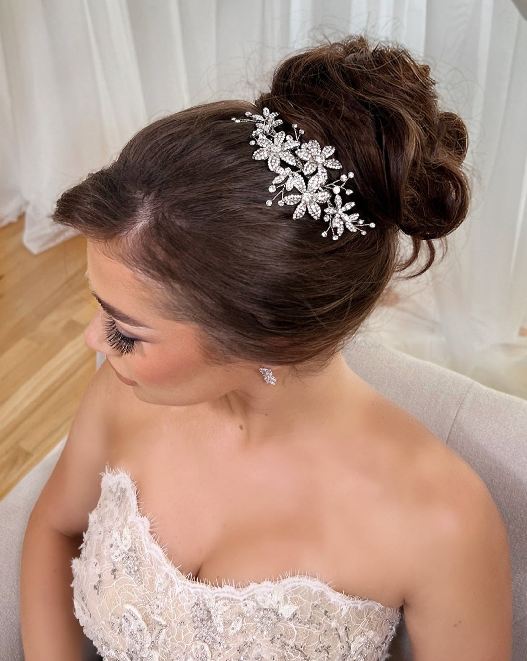 A bride with an elegant updo adorned with a Delicate Floral Comb (style no. 242692) sits on a cream sofa. She wears a strapless white lace gown and matching floral earrings, looking to her left against sheer curtains and wooden flooring.
