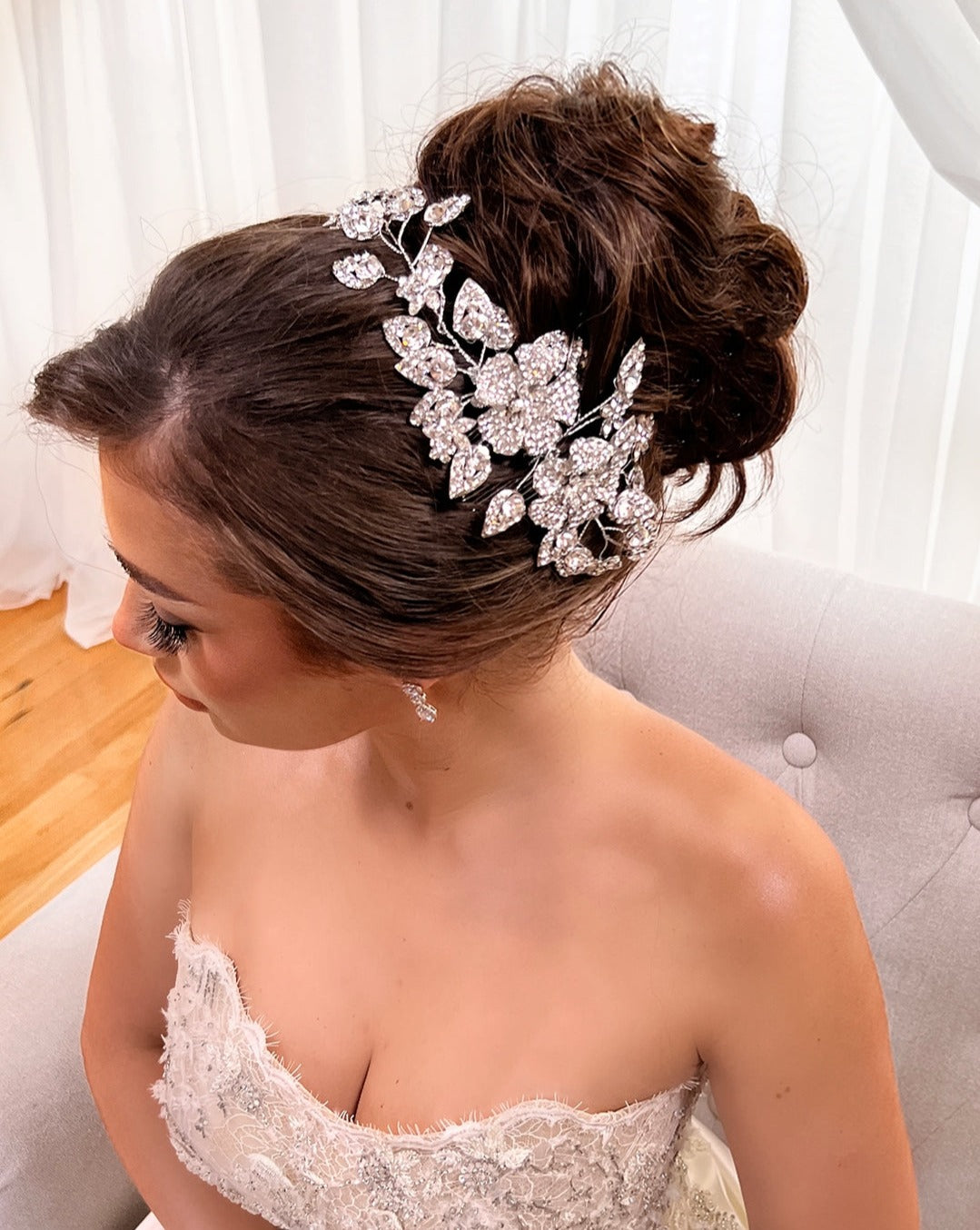 A woman in a strapless lace dress sits on a light gray chair. Her hair, styled in an elegant bun, is adorned with the Swarovski Crystal Flower & Leaflet Bridal Vine Style no. 80014. She gazes down, her long eyelashes highlighting her delicate earrings.