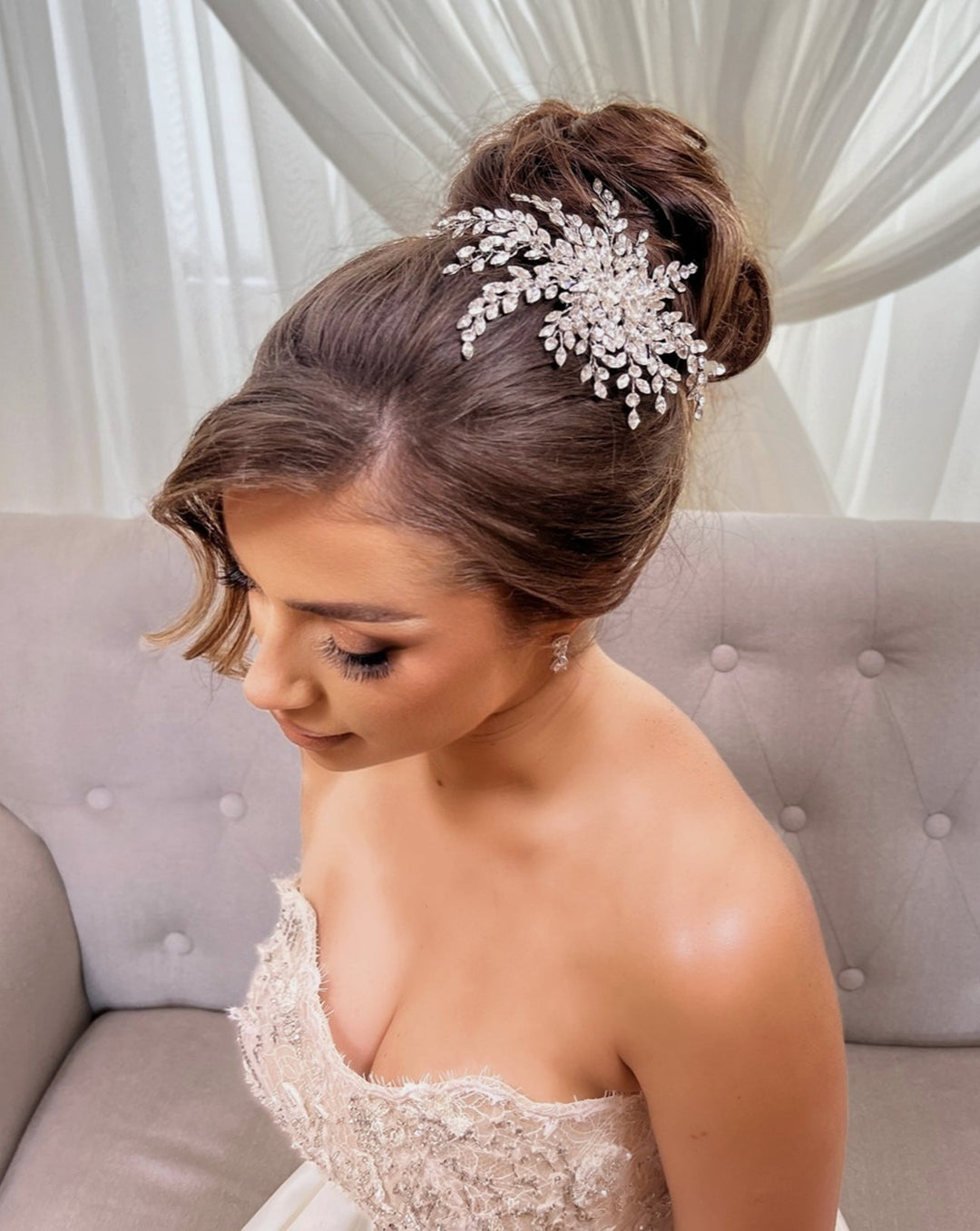 A woman with an intricate updo adorned by the Wispy Crystal Spray Comb no. 242672 wears a strapless lacy white dress, seated on a light beige sofa with sheer curtains in the background.