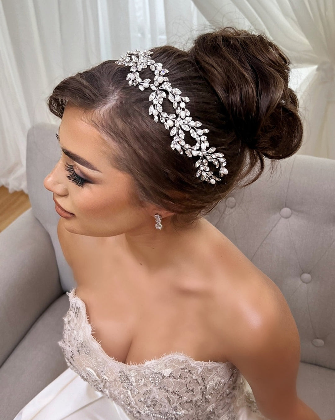 A bride wearing the Classic Pearl and Crystal Looping Hair Vine style no. 242644 and an elegant updo sits on a gray chair, dressed in a strapless lace wedding gown.