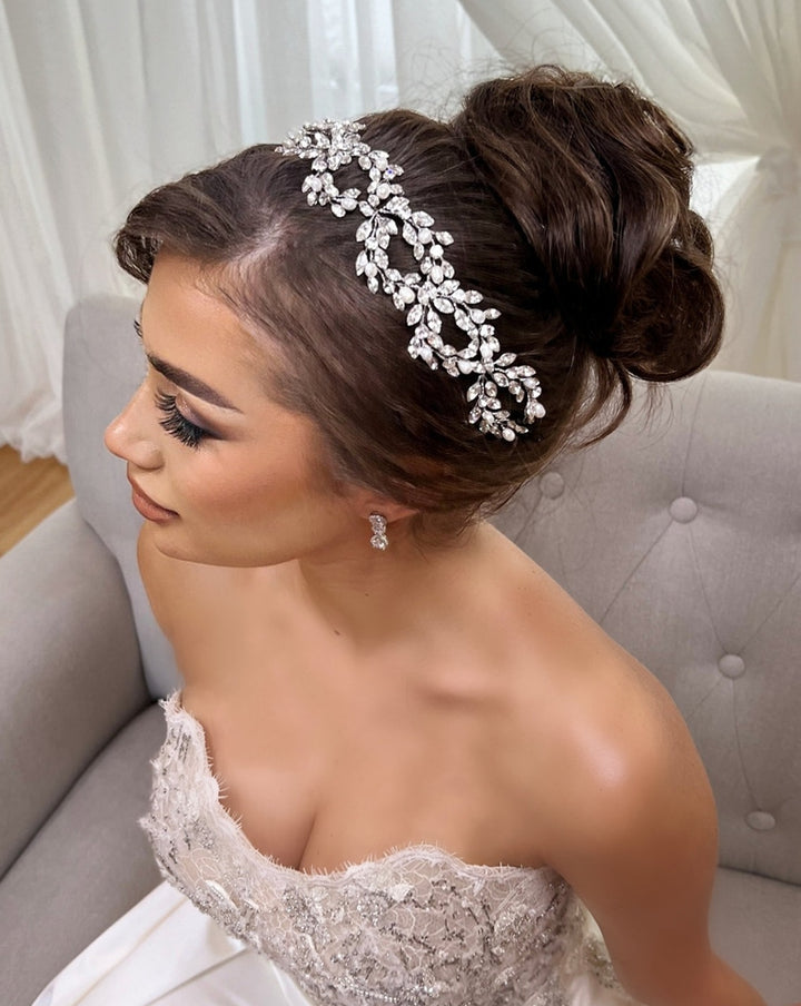 A bride wearing the Classic Pearl and Crystal Looping Hair Vine style no. 242644 and an elegant updo sits on a gray chair, dressed in a strapless lace wedding gown.
