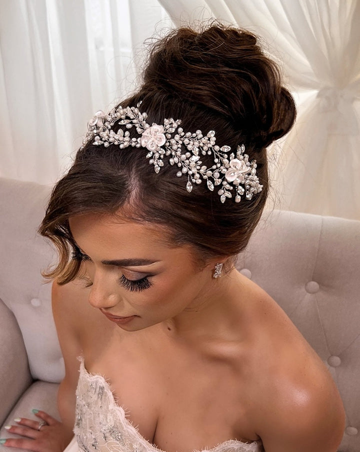 A bride with an elegant updo, accented by a Romantic Pearl and Porcelain Flower Swarovski Crystal Hair Vine (style no. 242714), sits on a cushioned chair. She wears a strapless lace wedding gown and has long, curled eyelashes. Light filters through sheer curtains in the background.
