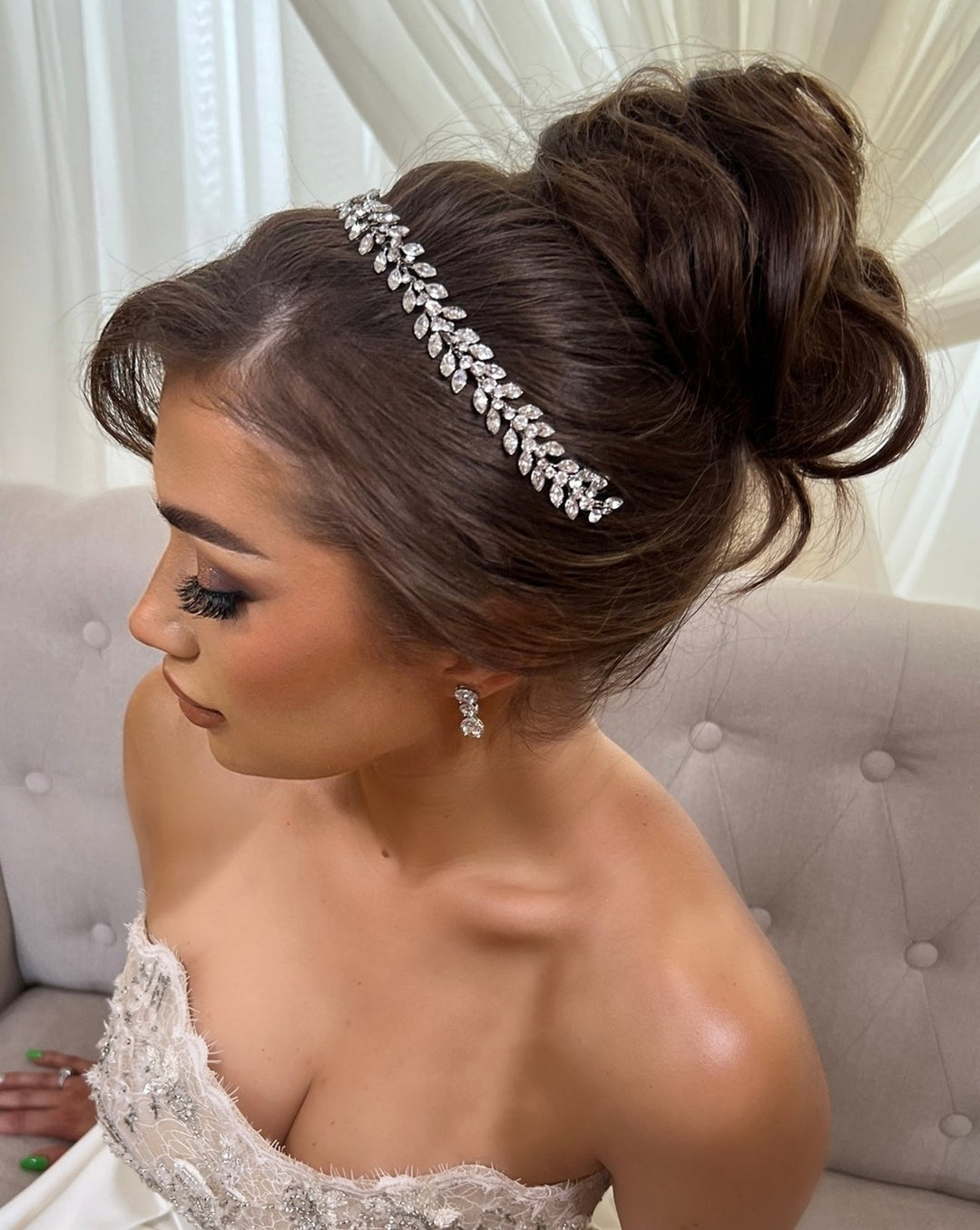 A woman with dark hair in an elegant bun, adorned with a Traditional Crystal Hair Vine crown (style no. 242710), sits on a light sofa. Shes wearing a strapless, lace-detailed gown and matching earrings, complemented by soft makeup and long lashes.