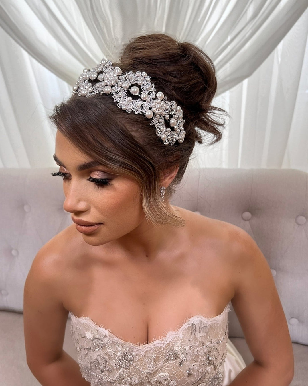 A woman in a strapless wedding dress with intricate beading sits on a beige couch. Her brown hair, styled in an updo, is adorned with the Glamorous Pearl and Crystal Headband (Style no. 392591). She has subtle makeup with long eyelashes and looks off to the side, set against white curtains.