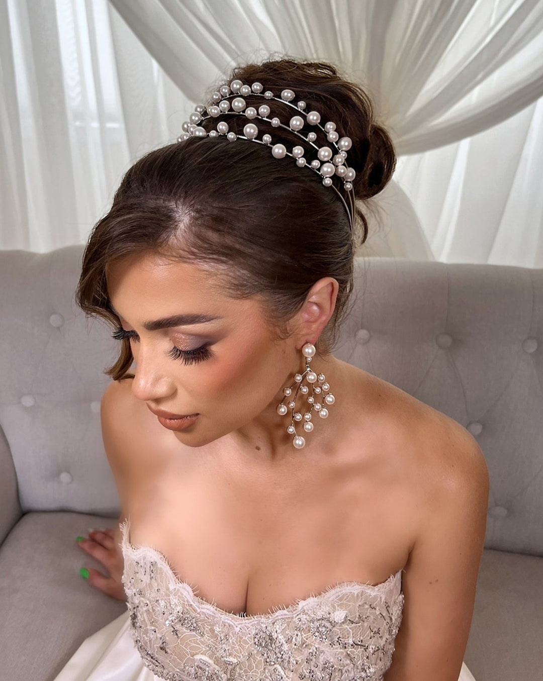 A woman in a strapless lace-detail dress sits on a gray tufted sofa. Her hair, styled in an updo, features Swarovski bridal combs and the Tiered Pearl Headband Style no. 80096. Large pearl earrings add to her elegance against sheer white curtains in the background.