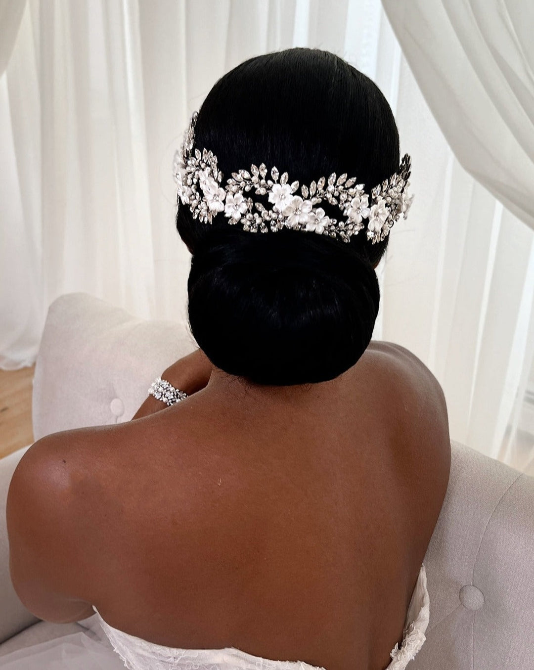 A person in a white strapless dress sits on a light-colored sofa with their back to the camera. Their hair is styled in an elegant bun, adorned with the Elegant Looping Flower Swarovski Crystal Hair Vine style no. 242727 . Sheer white curtains serve as the backdrop.