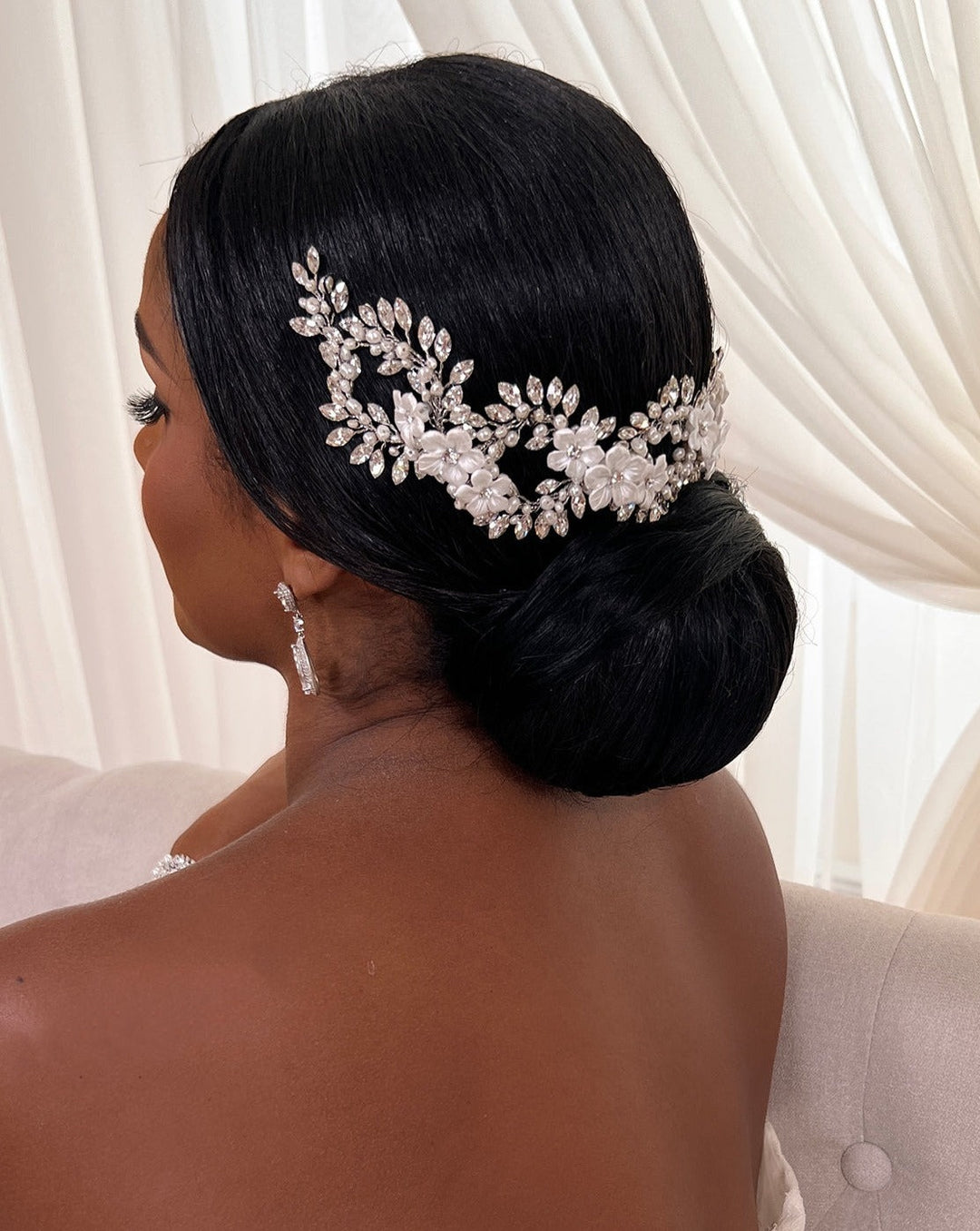 A woman with dark hair styled in an elegant low bun, adorned with an Elegant Looping Flower Wedding Hair Vine (style no. 242727) featuring Swarovski pearls and crystals. She wears delicate drop earrings and a strapless top against sheer white curtains in the background.
