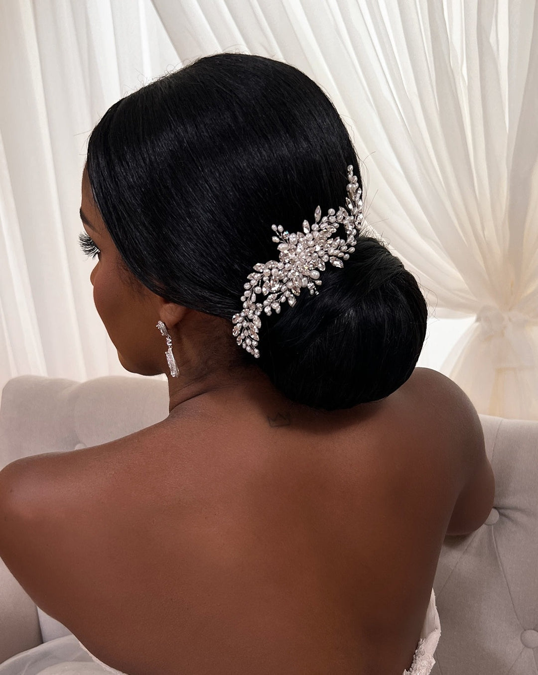 A person with dark hair styled in an elegant bun is adorned with the Flowing Cluster Hair Comb, style no. 242514, featuring pearl-like details and genuine crystals. They sit on a light-colored sofa with sheer, flowing curtains in the background.
